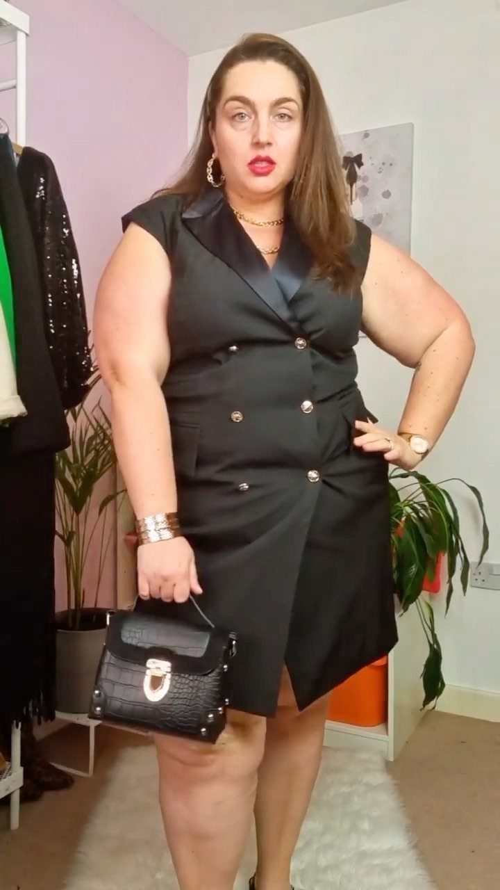 A woman posing in a black sleeveless satin-lapel waistcoat dress and holding a black handbag.
