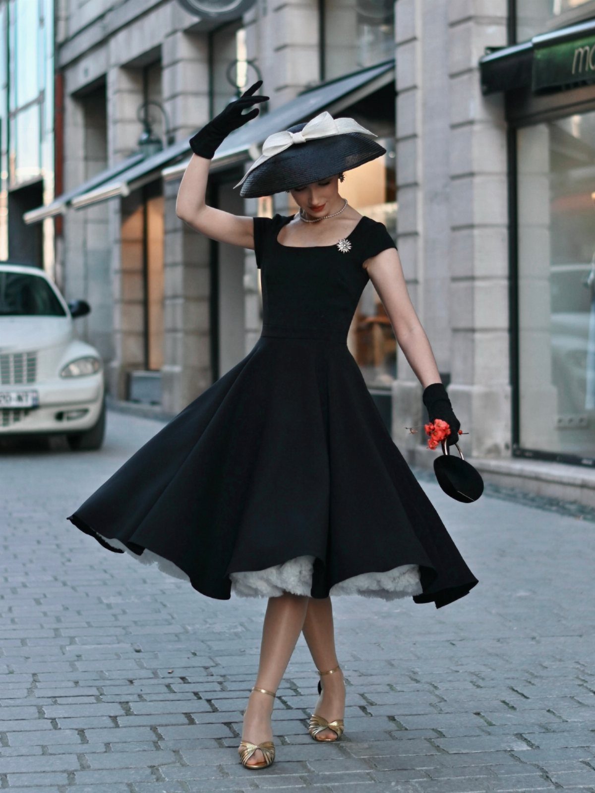 Woman in a black fit-and-flare dress and wide-brimmed hat posing on a cobblestone street.