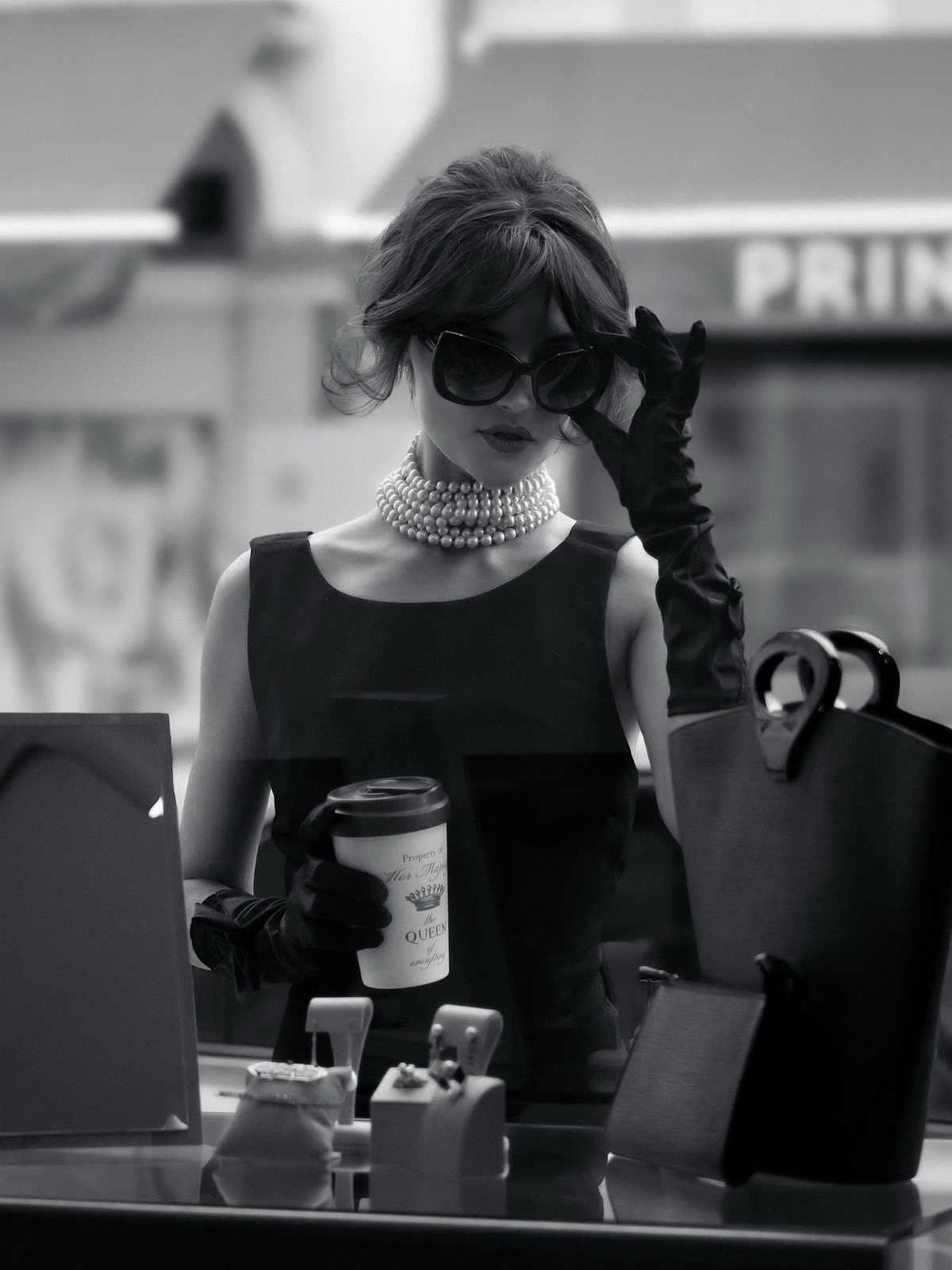 Woman in a black dress, pearl choker, and long gloves holding a coffee cup outdoors.