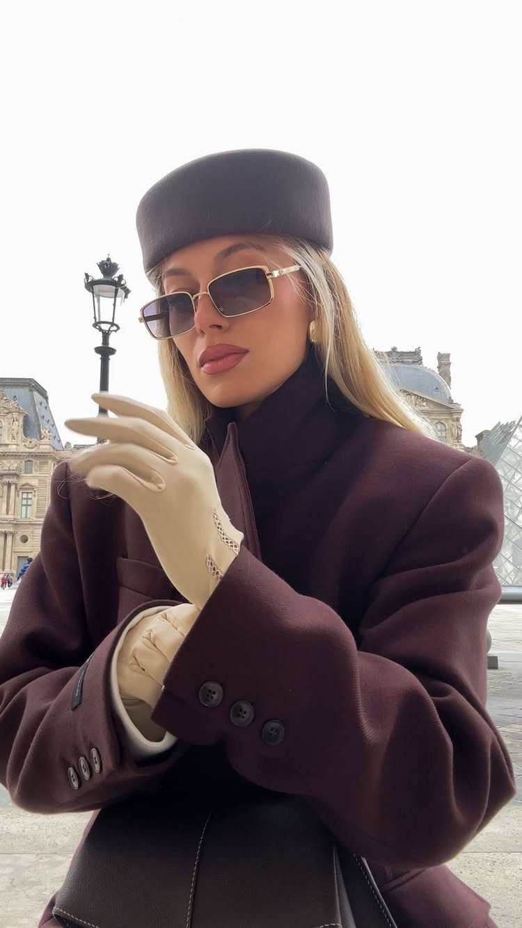 A woman in a burgundy coat, pillbox hat, and gloves posing outdoors.
