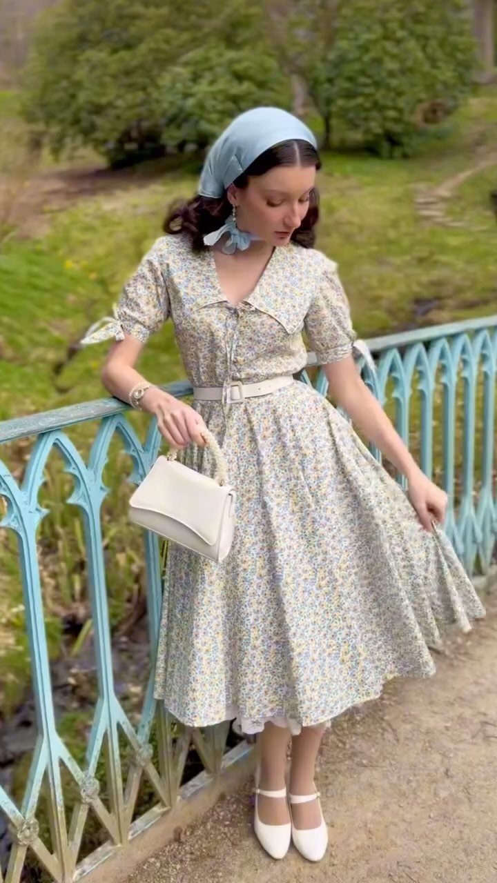 Woman in a floral tea dress and light blue headscarf standing by a blue railing.