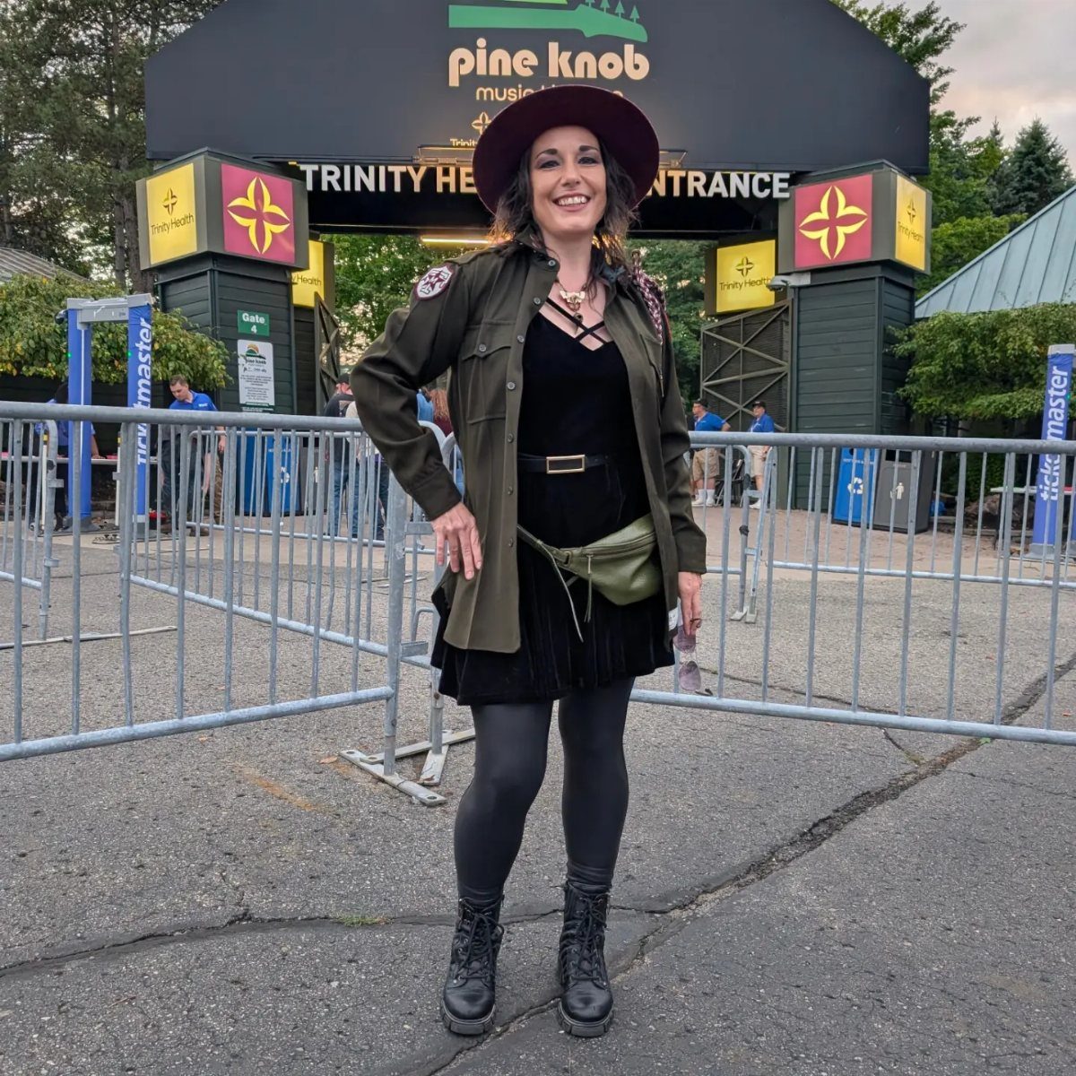 A smiling woman wearing an olive jacket, black skirt, and chunky combat boots standing outdoors.