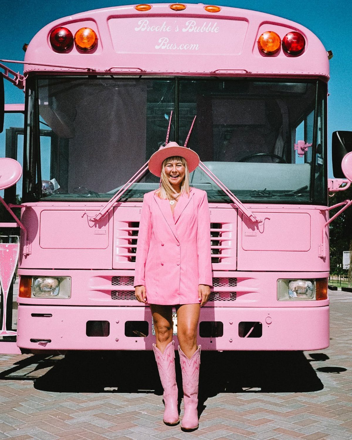 Woman in a pink blazer dress and cowboy boots standing in front of a pink bus.