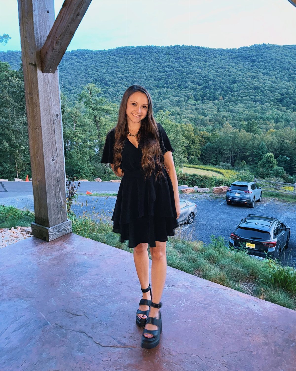 A smiling woman in a black dress and platform sandals standing on a stone patio.