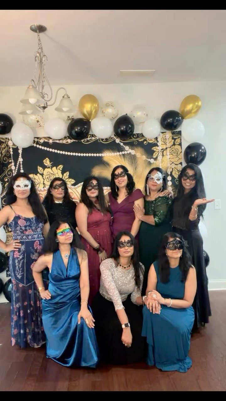 Group of women in elegant dresses and masquerade masks posing in front of a decorated backdrop.