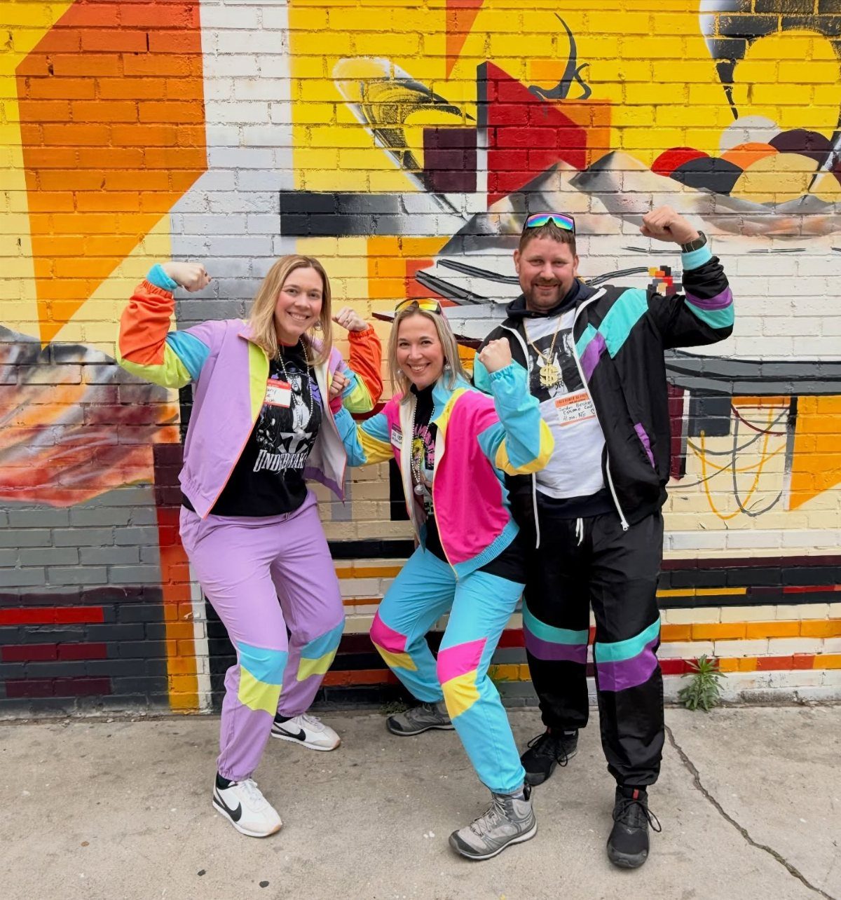 Three people posing in bright, neon-colored 80s-style windbreaker tracksuits against a colorful graffiti wall.