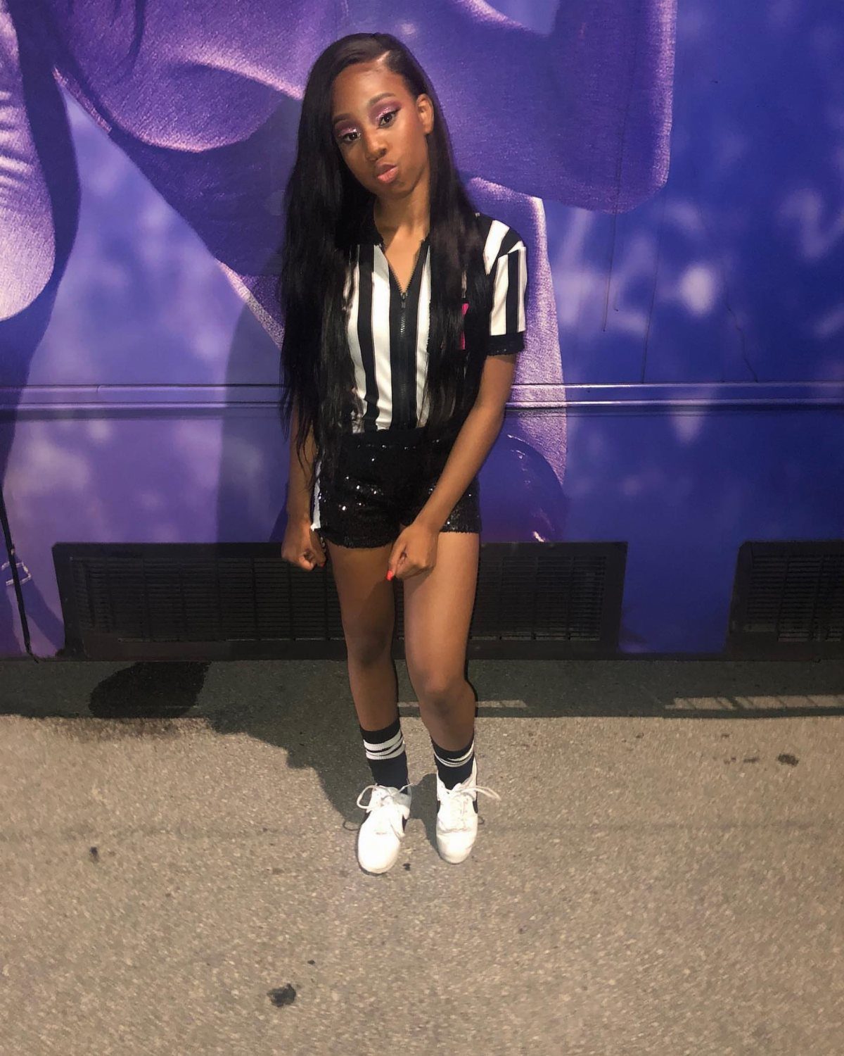 A woman in a striped referee shirt and sequined shorts posing against a purple wall.