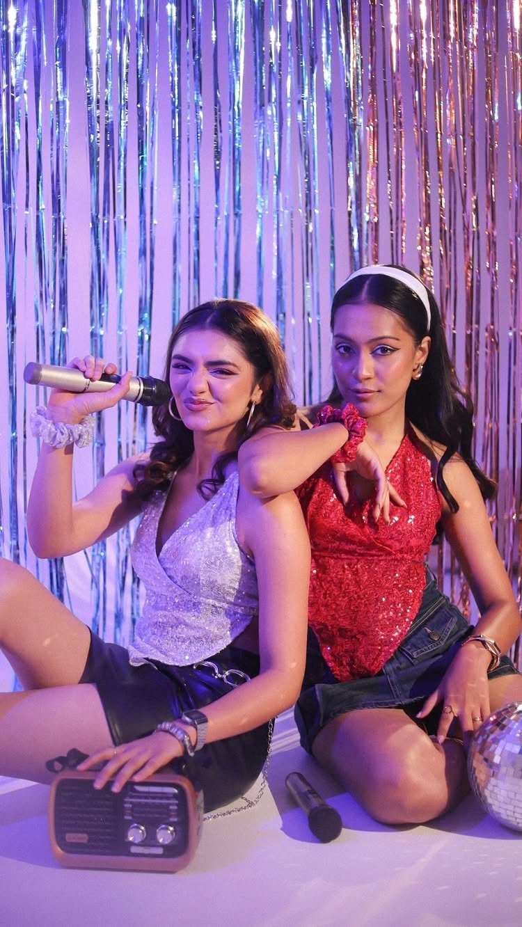 Two women in sparkly party outfits posing in front of a silver tinsel backdrop with microphones.