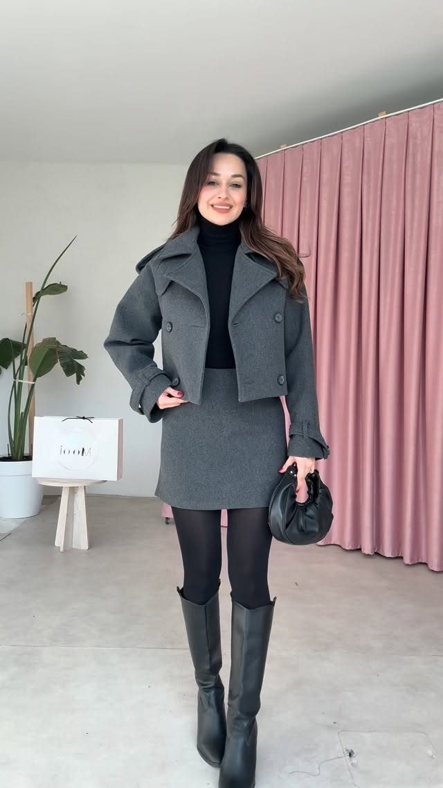A woman wearing a grey skirt suit, black turtleneck, and black boots posing indoors.