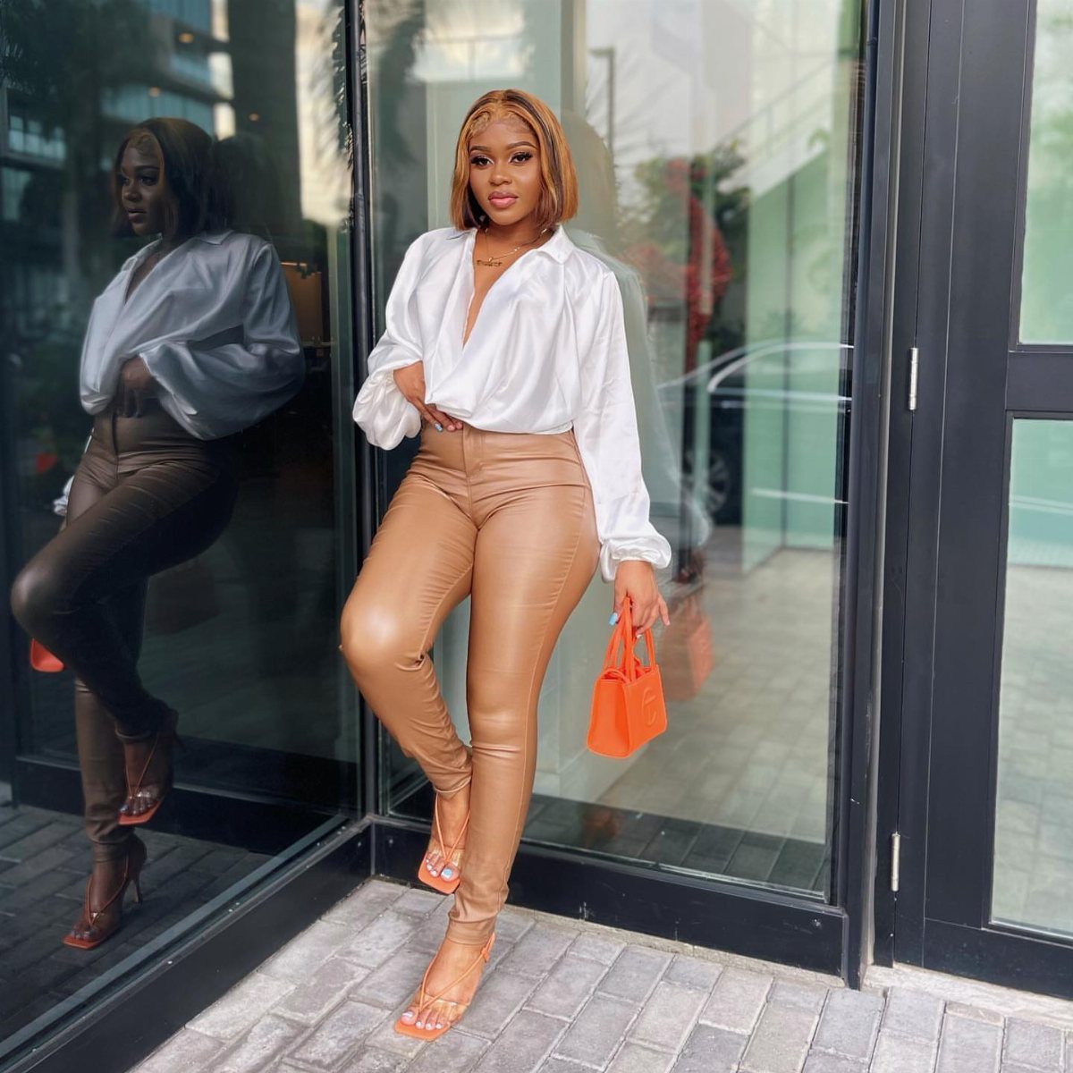 A woman posing in tan faux leather pants, a white blouse, and orange heels.