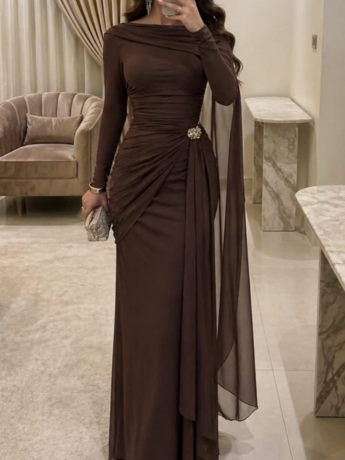 Woman in a long-sleeved brown draped evening gown with a jeweled brooch and sheer cape.