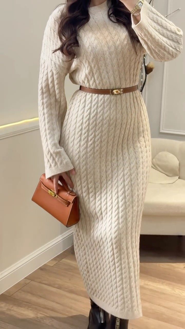 A person wearing a long cream cable-knit sweater dress with a brown belt and handbag.