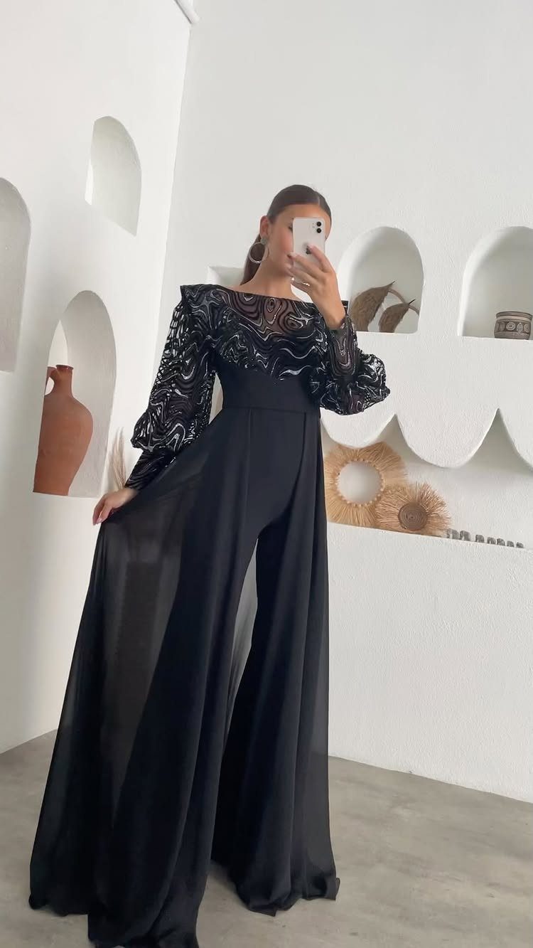 A woman wearing a black sequined wide-leg jumpsuit posing in a white room.