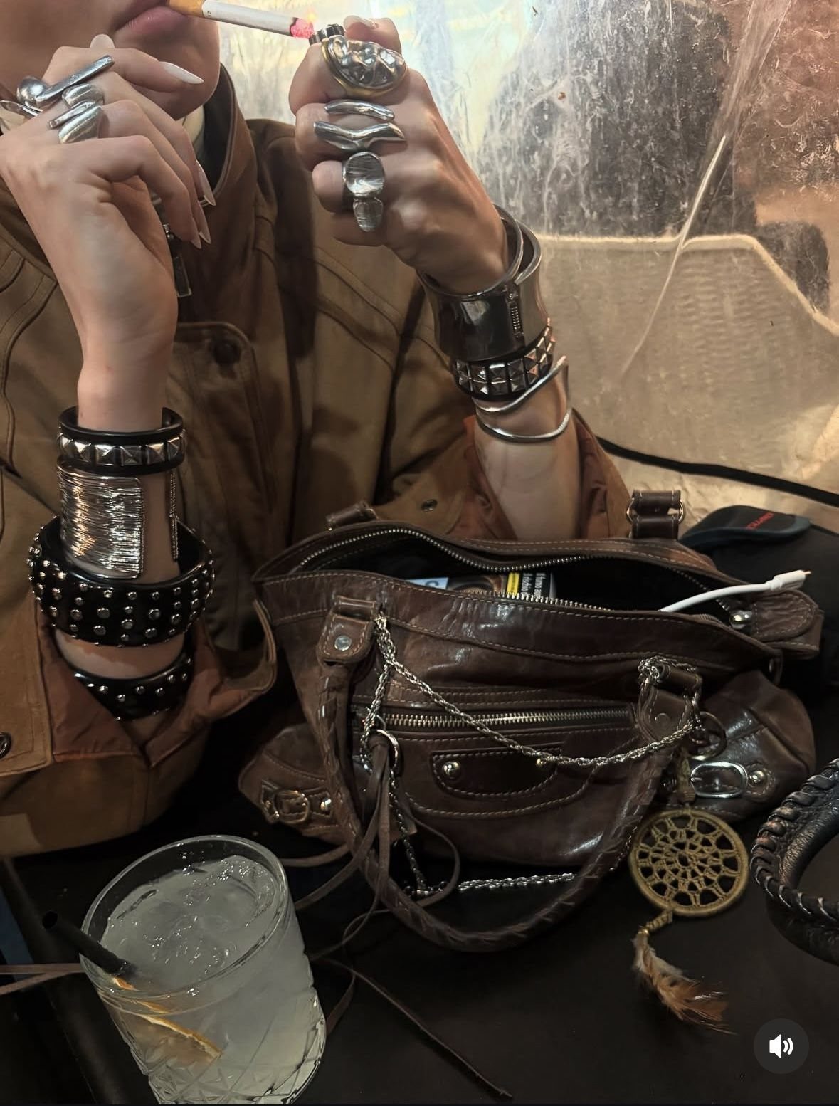 Person wearing large silver rings and multiple studded leather bracelets while holding a cigarette.