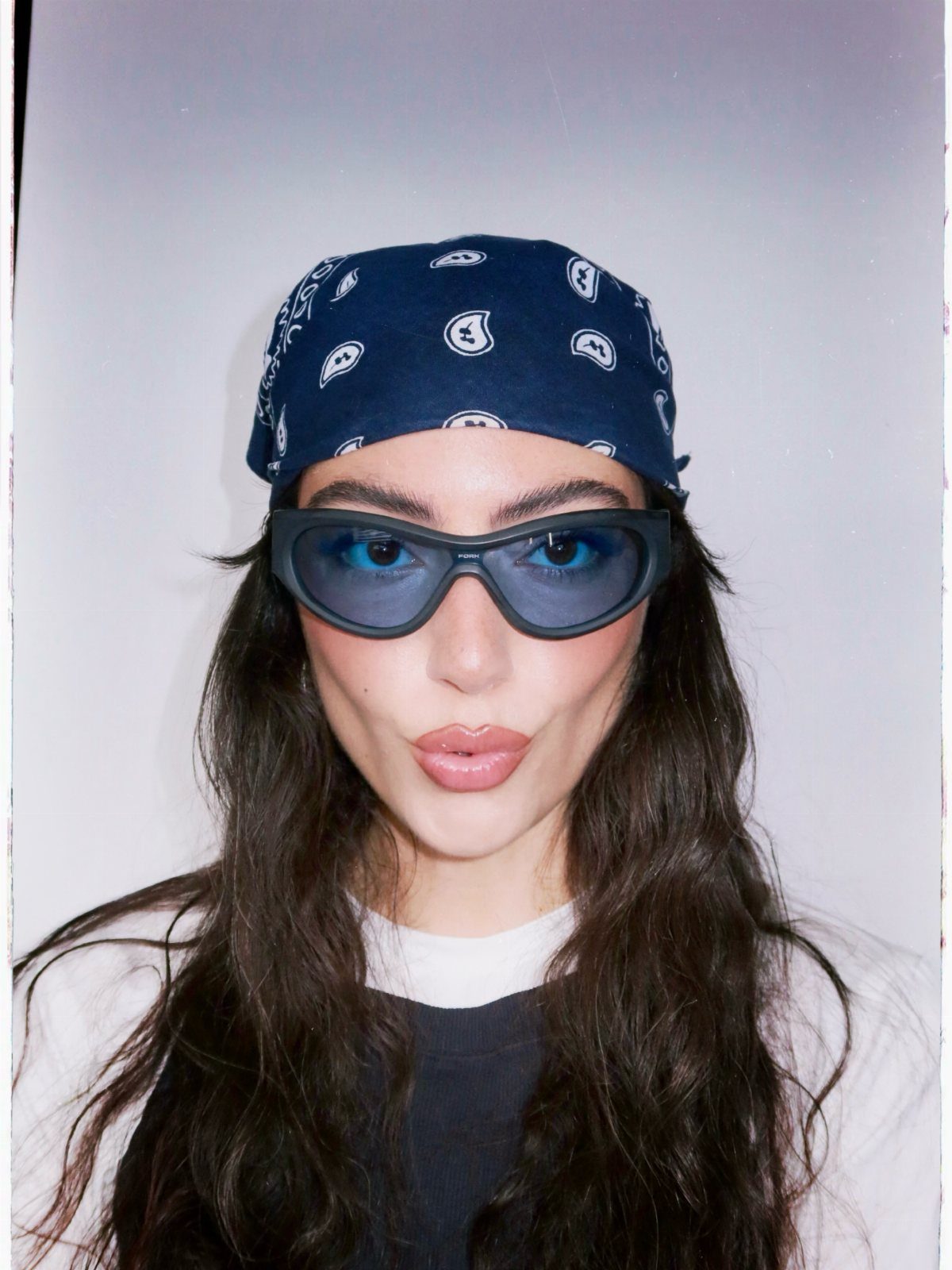 A woman wearing a blue bandana and large blue-tinted sunglasses against a light background.
