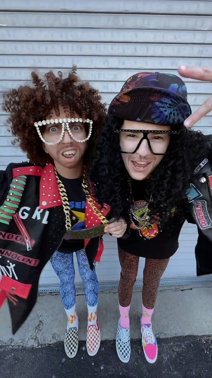 Two people wearing flamboyant LMFAO-inspired costumes with large wigs and oversized glasses.