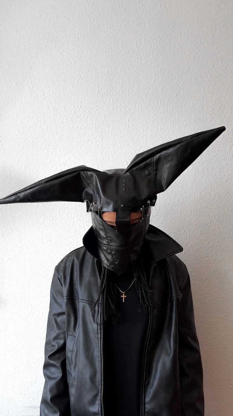 A person wearing a dramatic black leather mask with large pointed wing-like extensions and a leather jacket.