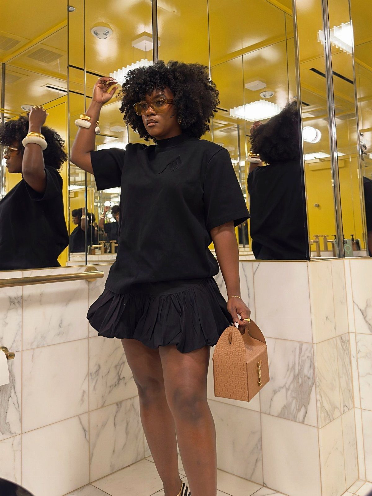 Woman wearing a black t-shirt and a black pleated mini skirt holding a tan handbag.