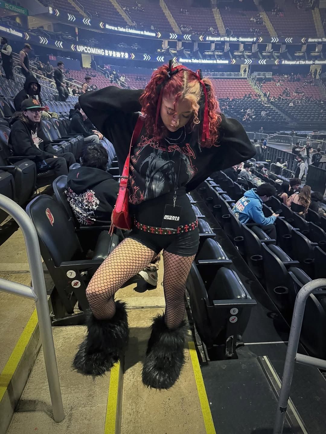 Person with red hair wearing a black oversized graphic hoodie and fishnet tights in a stadium.