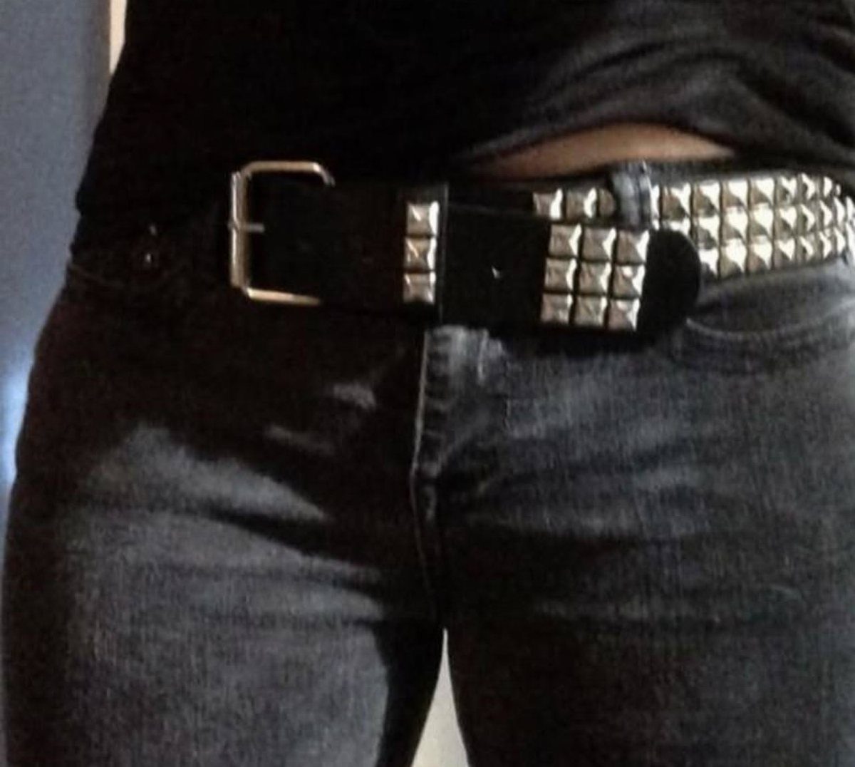 A wide black leather belt with large silver square studs on dark denim jeans.