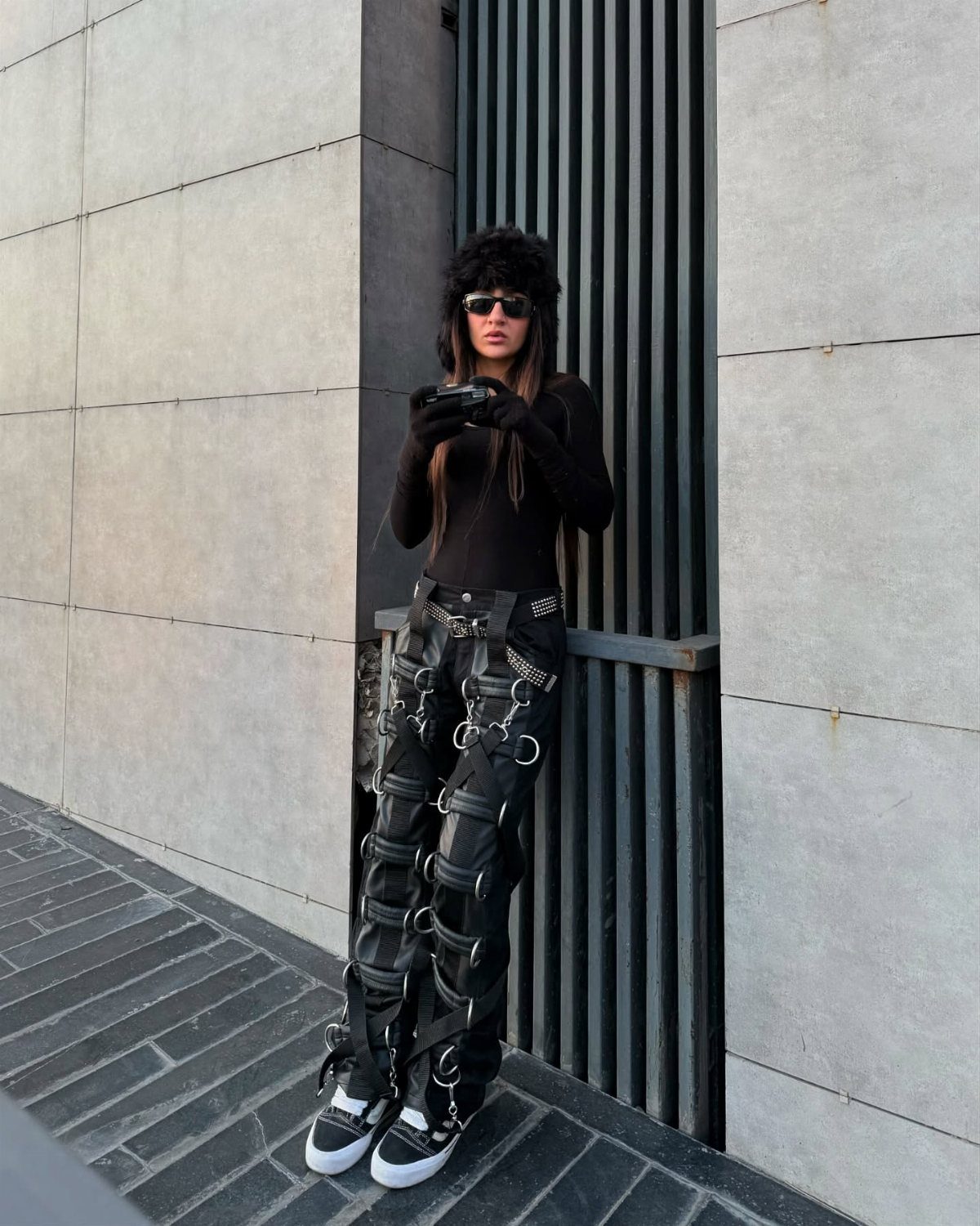 A person wearing black sunglasses and intricate black bondage-style trousers with silver rings and straps.