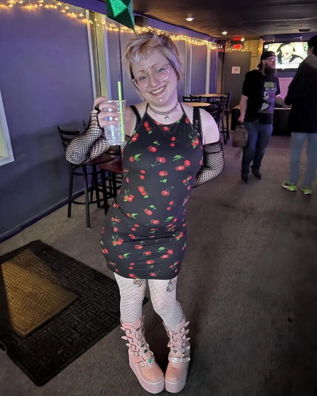 A woman in a cherry print dress and platform boots smiling in a club.