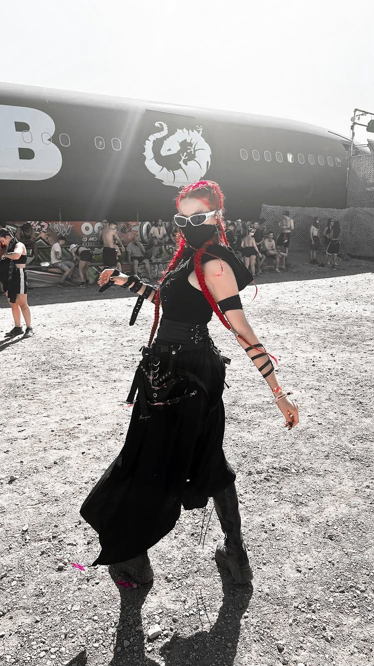 A person posing in a black modest rave outfit with red hair and sunglasses outdoors.