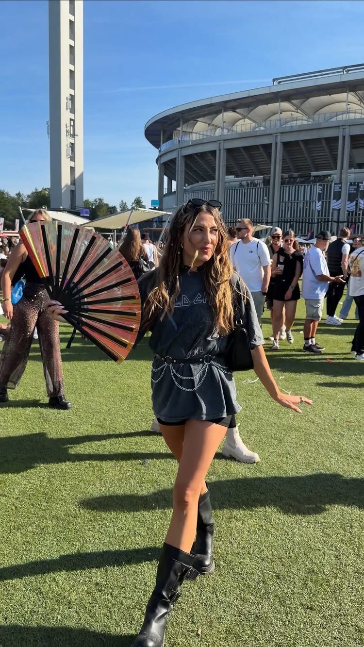 A woman in a dark oversized t-shirt and black boots holding a large decorative fan outdoors.