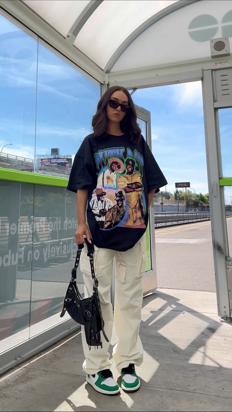 Woman in oversized graphic t-shirt and white cargo pants standing by a bus shelter.
