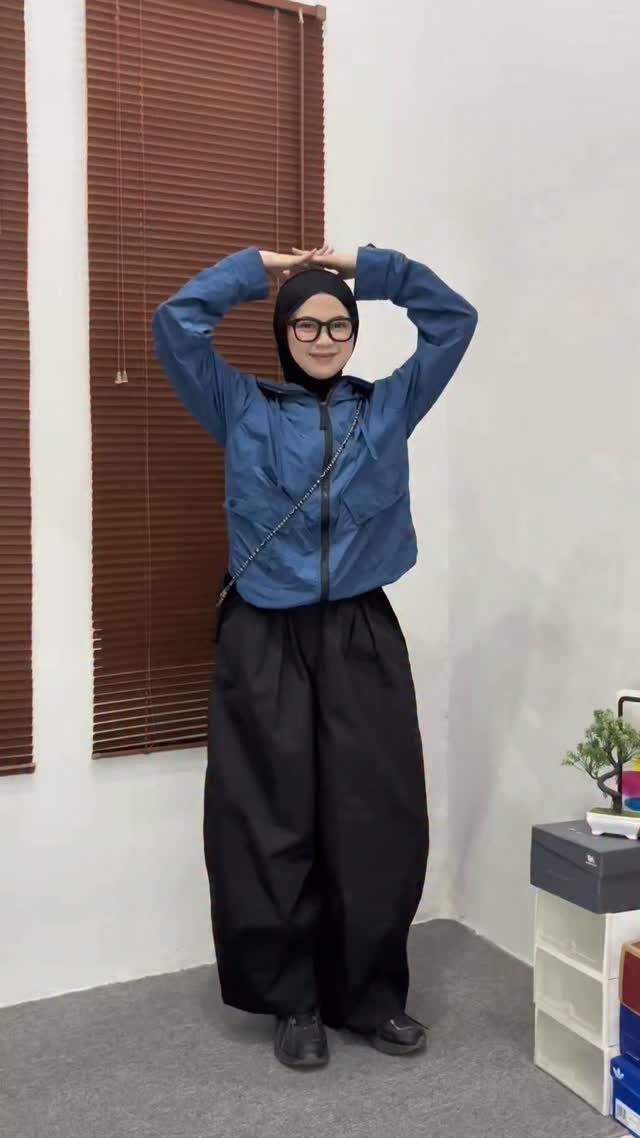 Person posing in a blue windbreaker jacket and voluminous black trousers against a white wall.