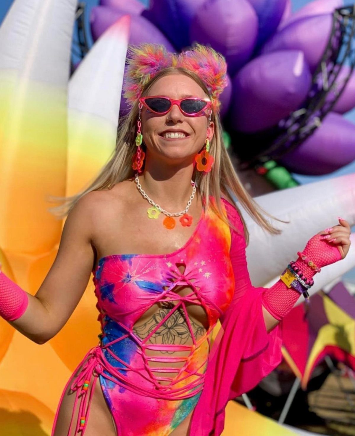 Smiling woman wearing colorful rave accessories including pink sunglasses, flower earrings, and a beaded necklace.