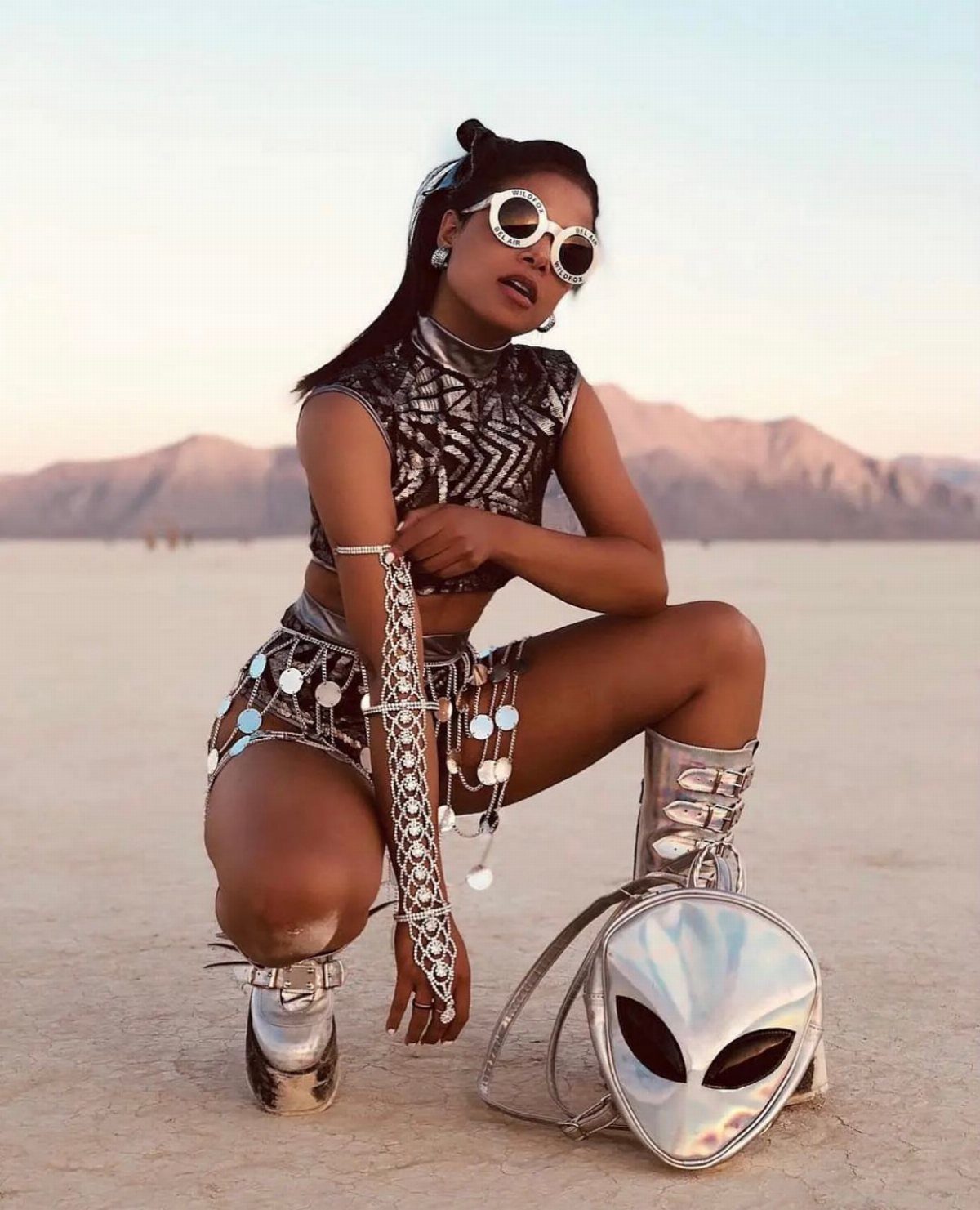 Woman in silver metallic festival outfit crouching in a desert landscape with mountains.