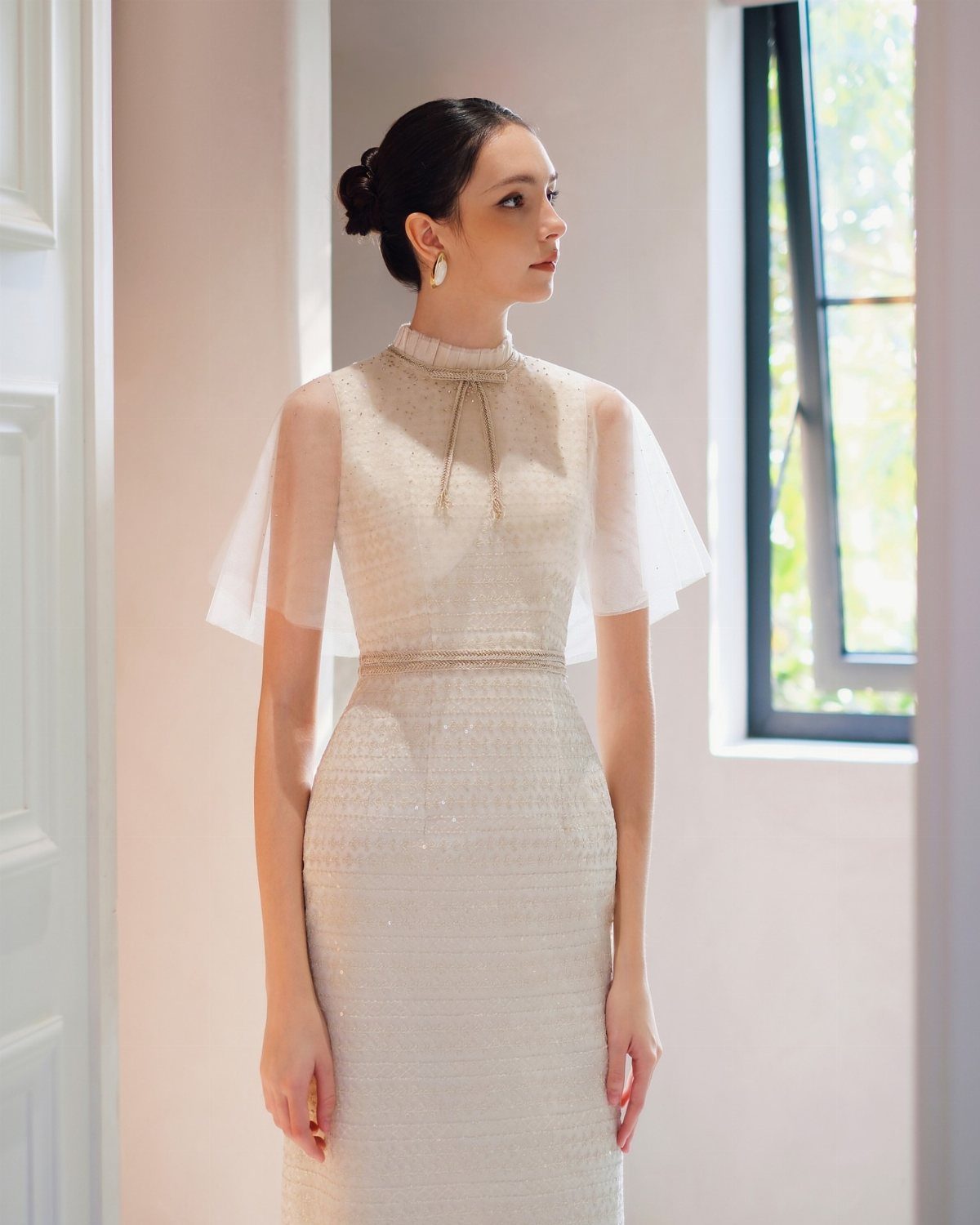 A woman in a formal cream sequined gown with a sheer tulle capelet standing indoors.