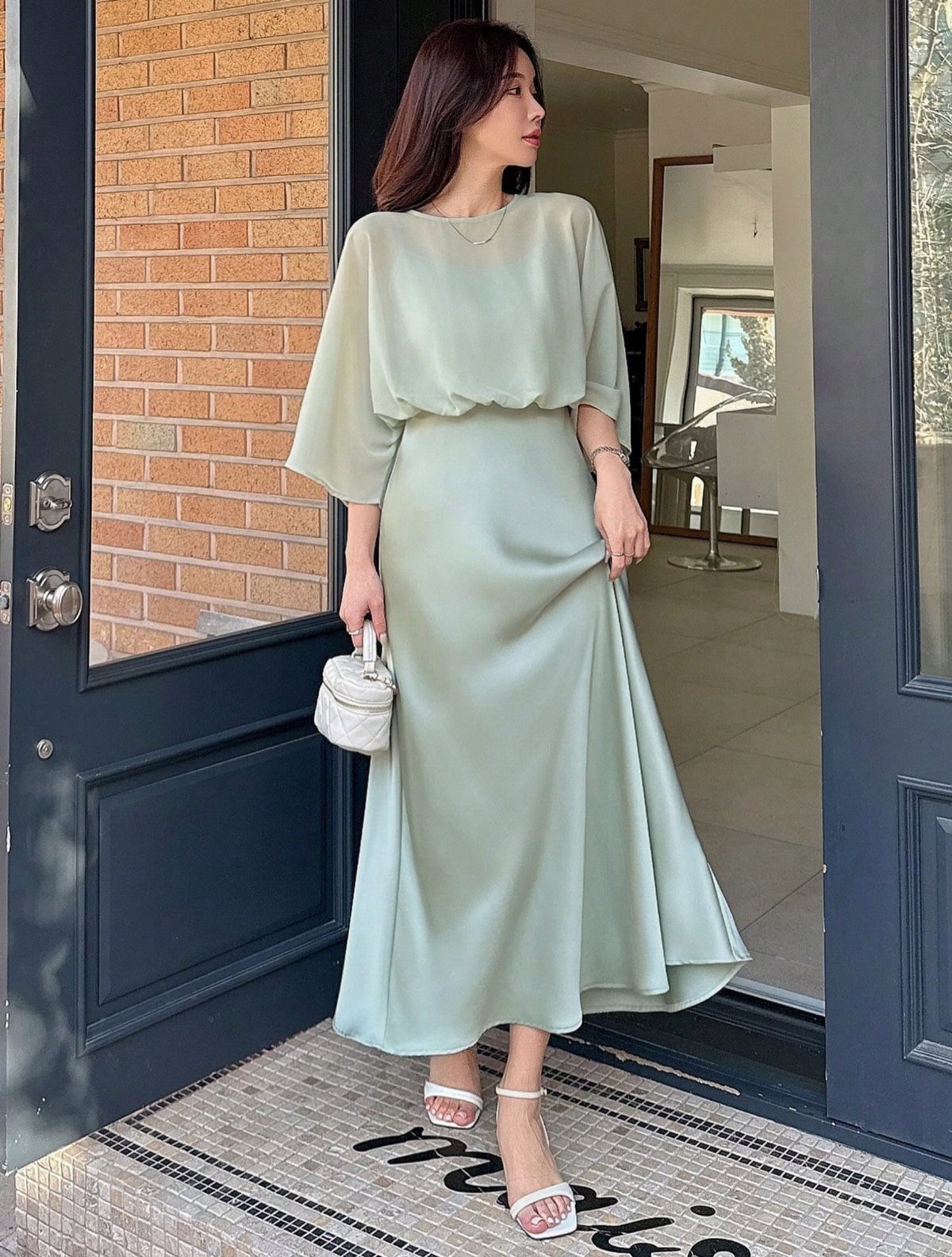 A woman posing in a sage green satin midi dress with wide sleeves and white sandals.