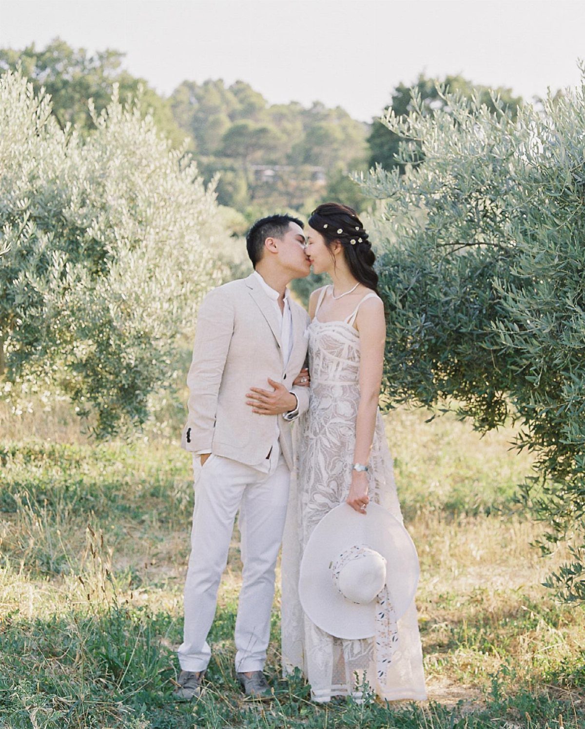 A man in a beige suit and a woman in a white lace dress kissing outdoors.