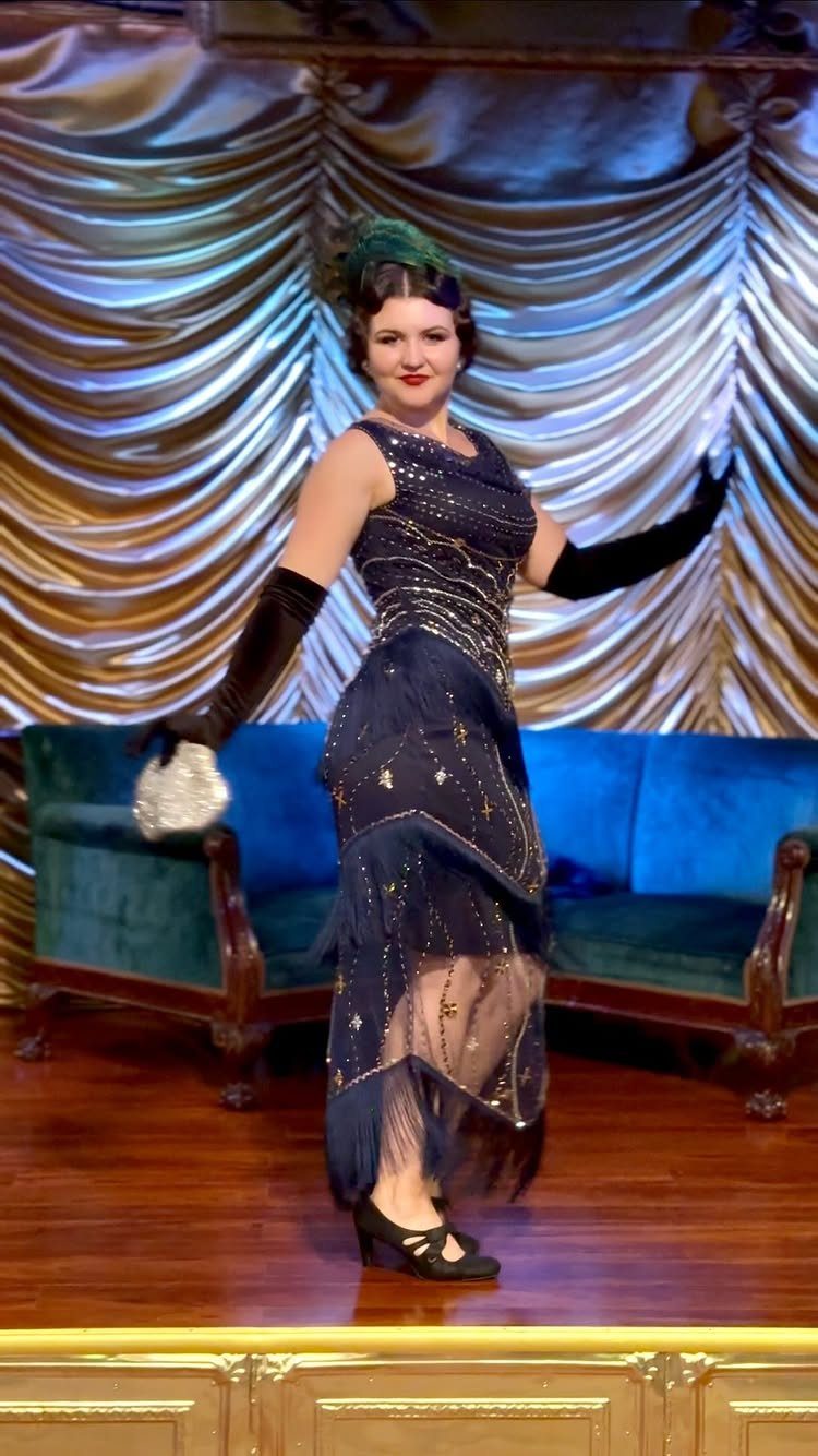Woman in a dark blue embellished flapper dress posing against gold curtains and a blue sofa.