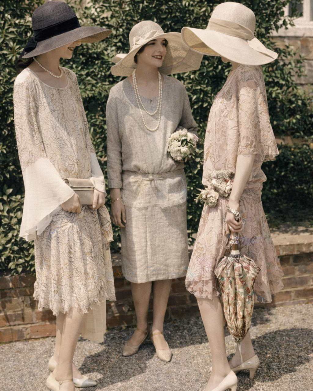Three women in vintage lace and embroidered dresses wearing wide-brimmed hats outdoors.