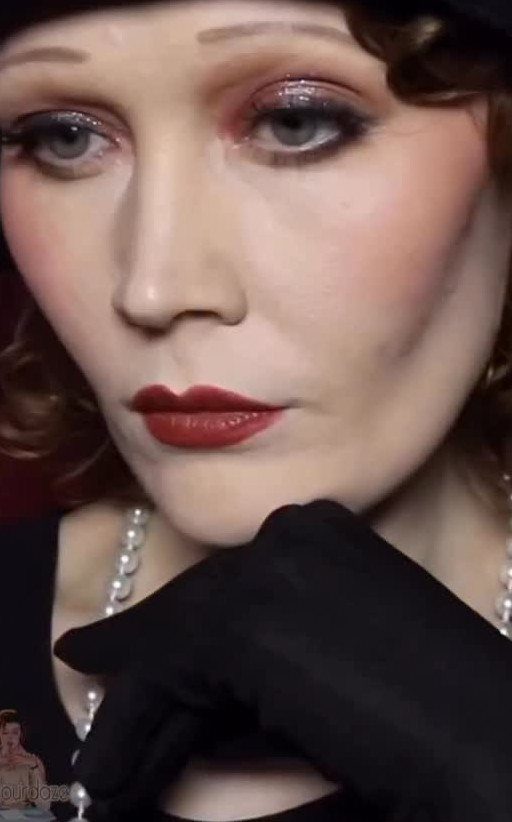 A woman wearing a dark cloche hat, smoky eye makeup, and red lipstick.