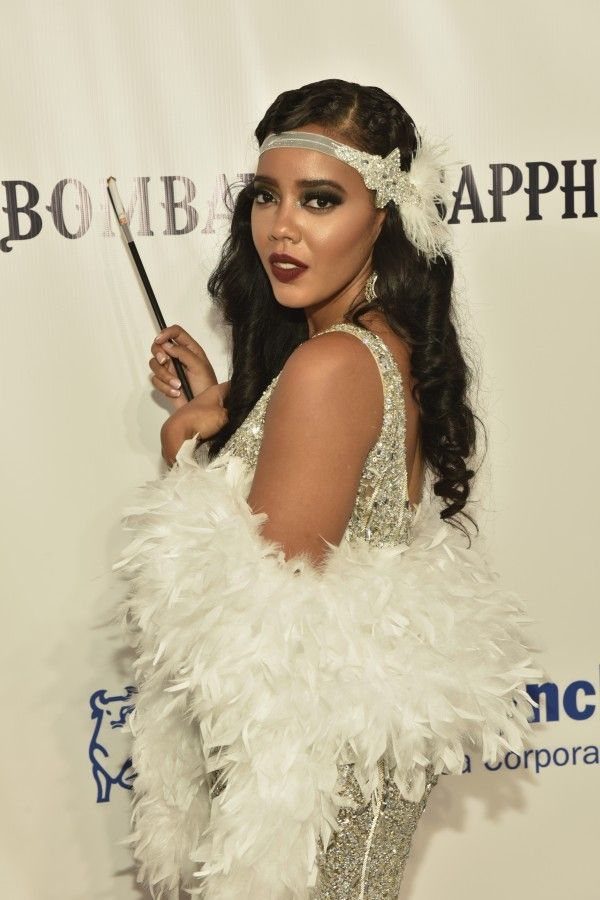 Woman in a sequined dress and white feather boa posing against a branded backdrop.