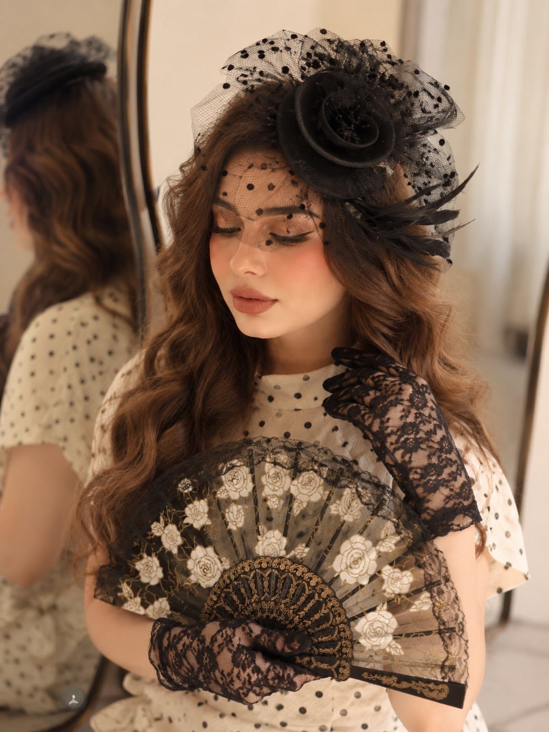 Woman with long wavy hair wearing black lace gloves and holding an ornate folding fan.