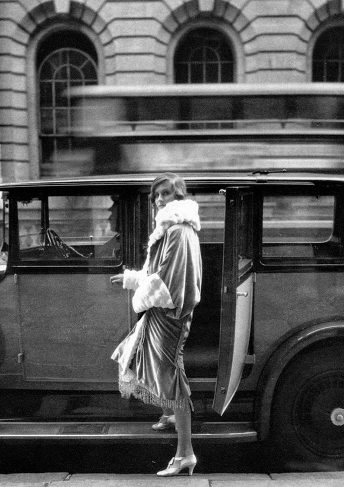 A woman wearing a long coat with a white fur collar stands next to a vintage car.