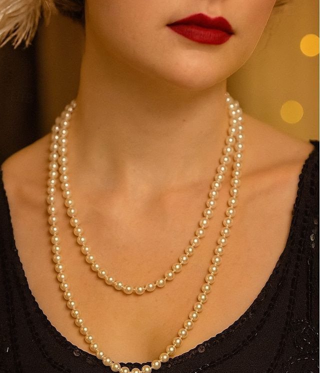 A person wearing a long double-strand pearl necklace and red lipstick against a blurred background.