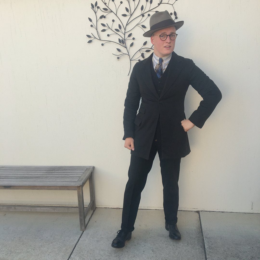 A man posing in a dark three-piece suit, patterned tie, and grey fedora hat.