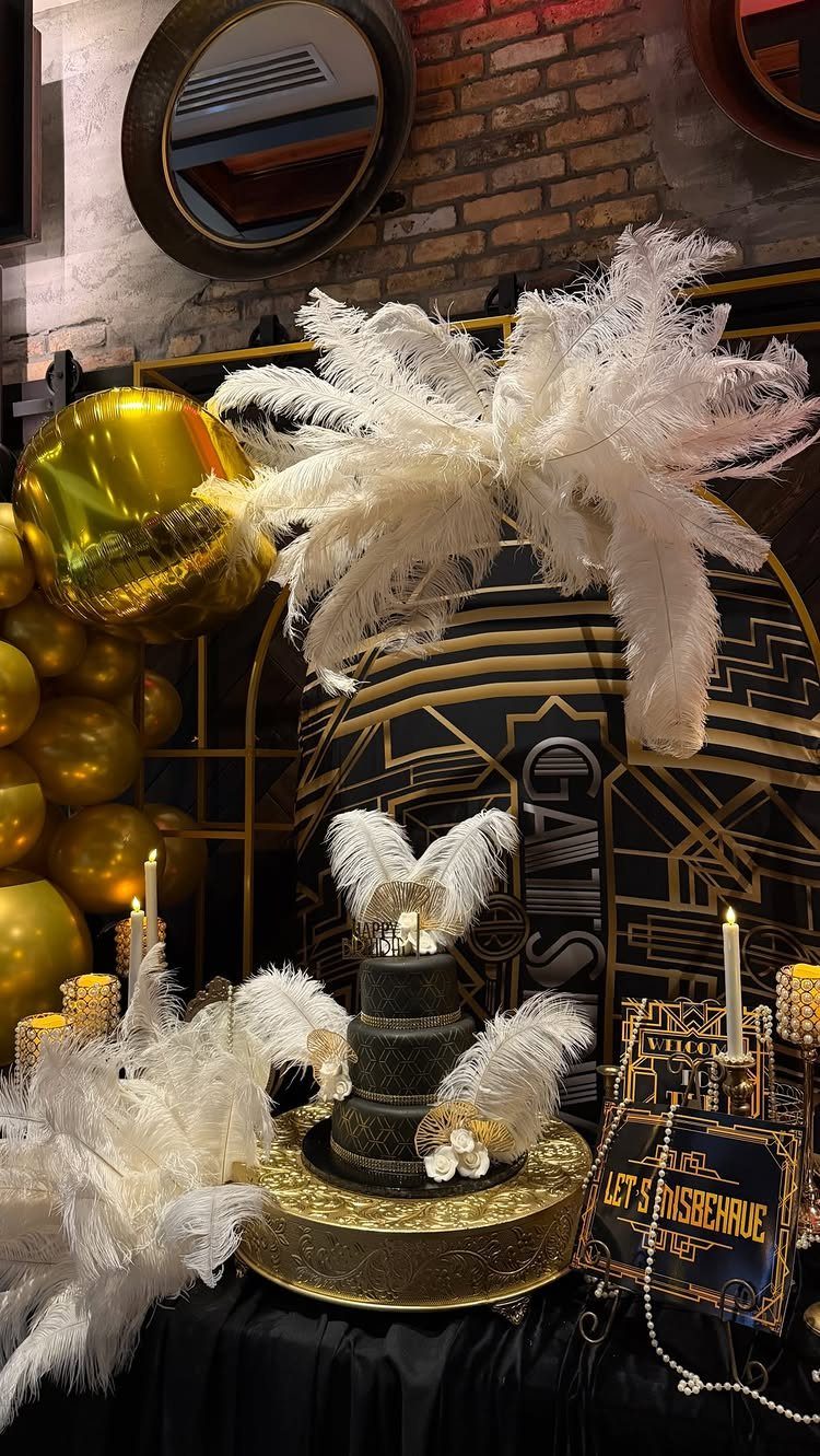 A black tiered cake decorated with white feathers sits on a gold stand amidst gold balloons.