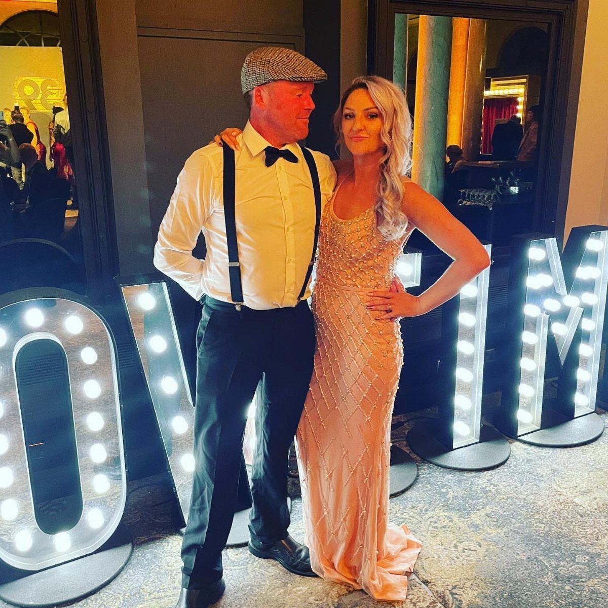 A man in 1920s attire and a woman in a beaded gown posing by marquee letters.