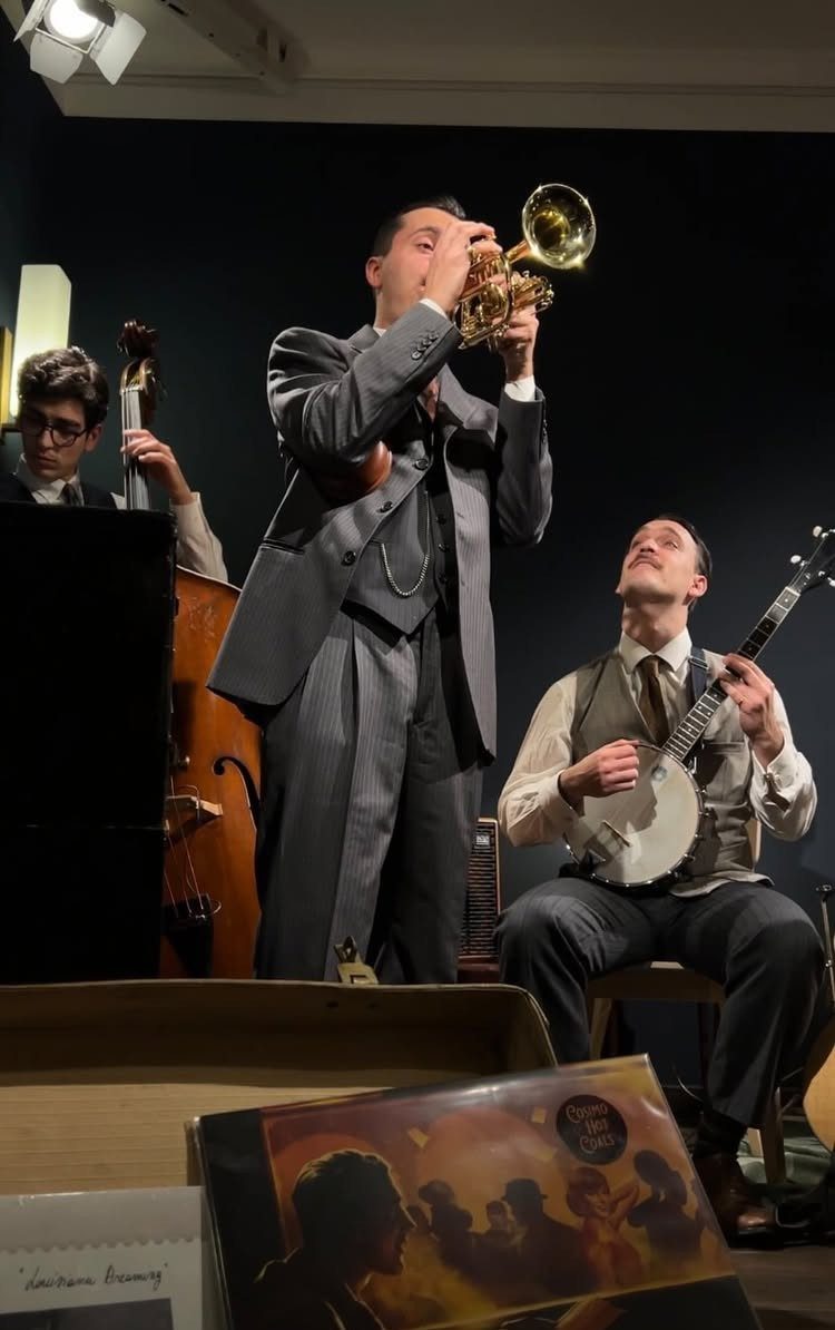 Three men in vintage clothing playing trumpet, banjo, and double bass on a stage.