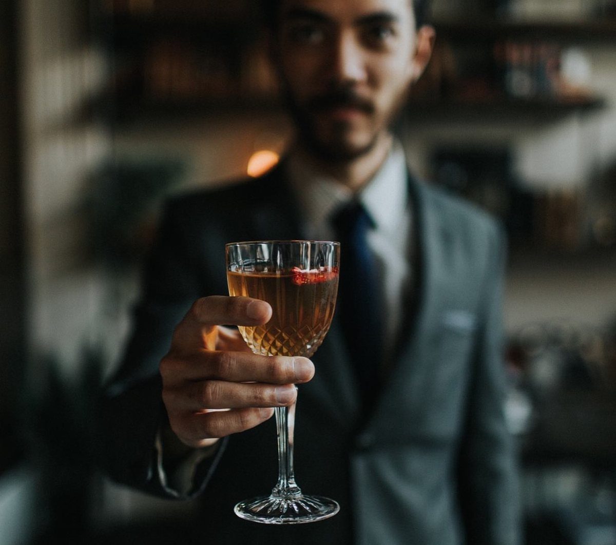 A man in a suit holding out a cocktail glass filled with an amber drink.