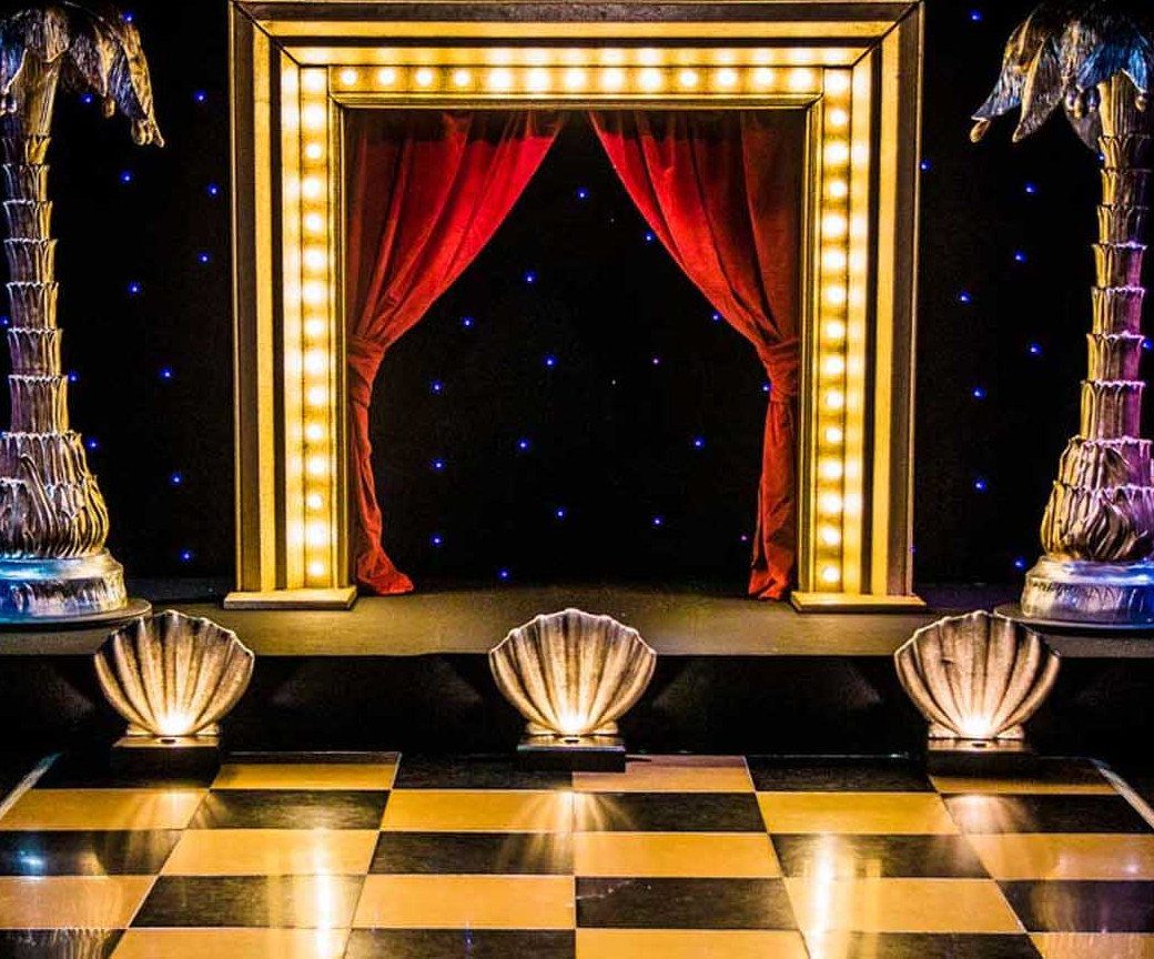 A stage with red curtains, marquee lights, and a checkered dance floor.