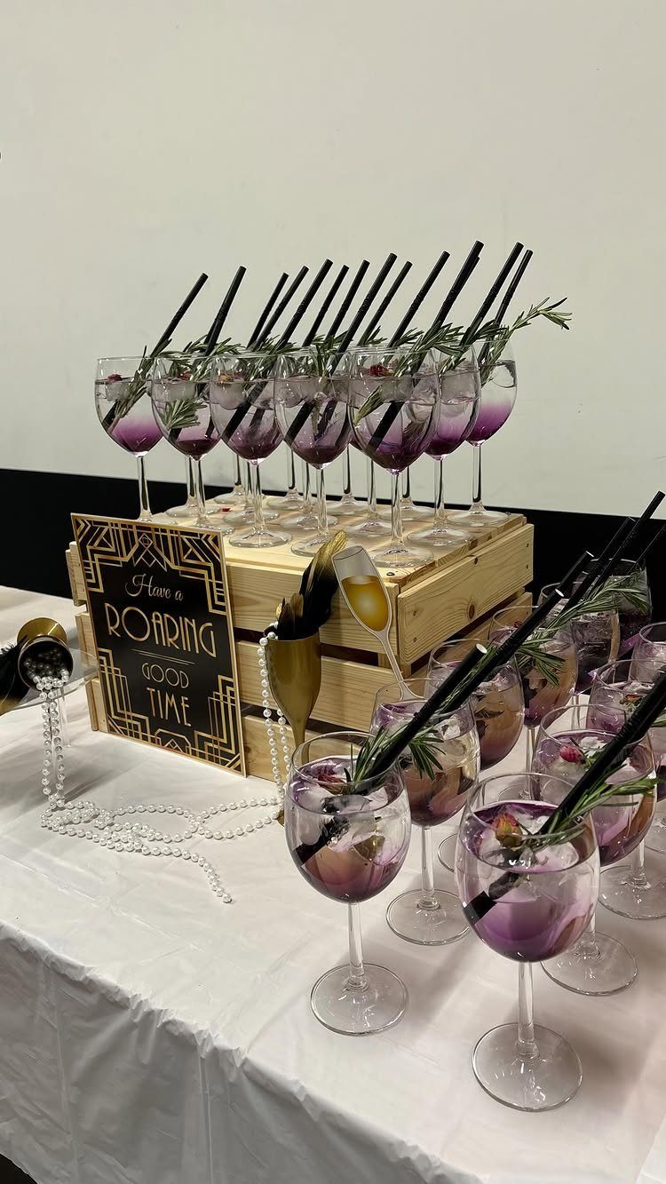 Cocktail glasses with purple drinks and rosemary garnishes arranged on a wooden crate and white tablecloth.