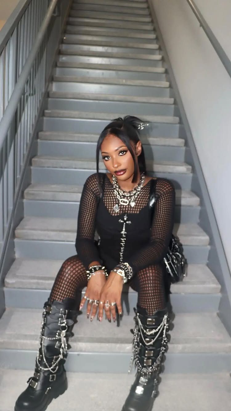 Woman in all-black mesh outfit and chain-adorned combat boots sitting on stairs.